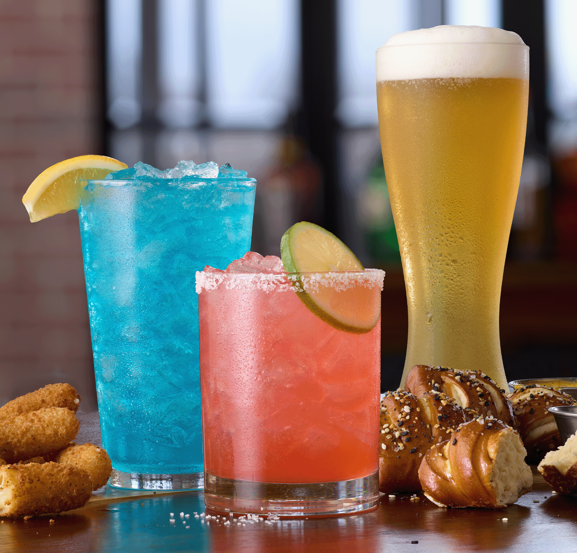  A blue cocktail with a lemon, a pink cocktail with a lime, and a glass of beer with soft pretzels on the table