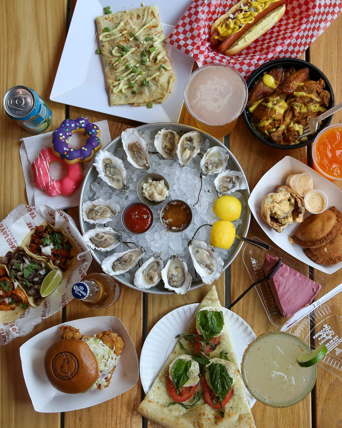 A top down view of many foods from different vendors on a table, including seafood, pizza, a burger, donuts, drinks, tacos, a hot dog, and cake