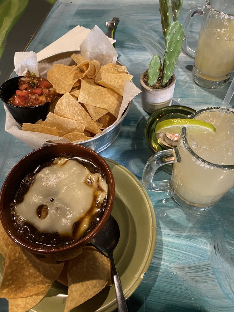 A basket of chips and salsa, a bowl of soup, and two margaritas, with a potted cactus