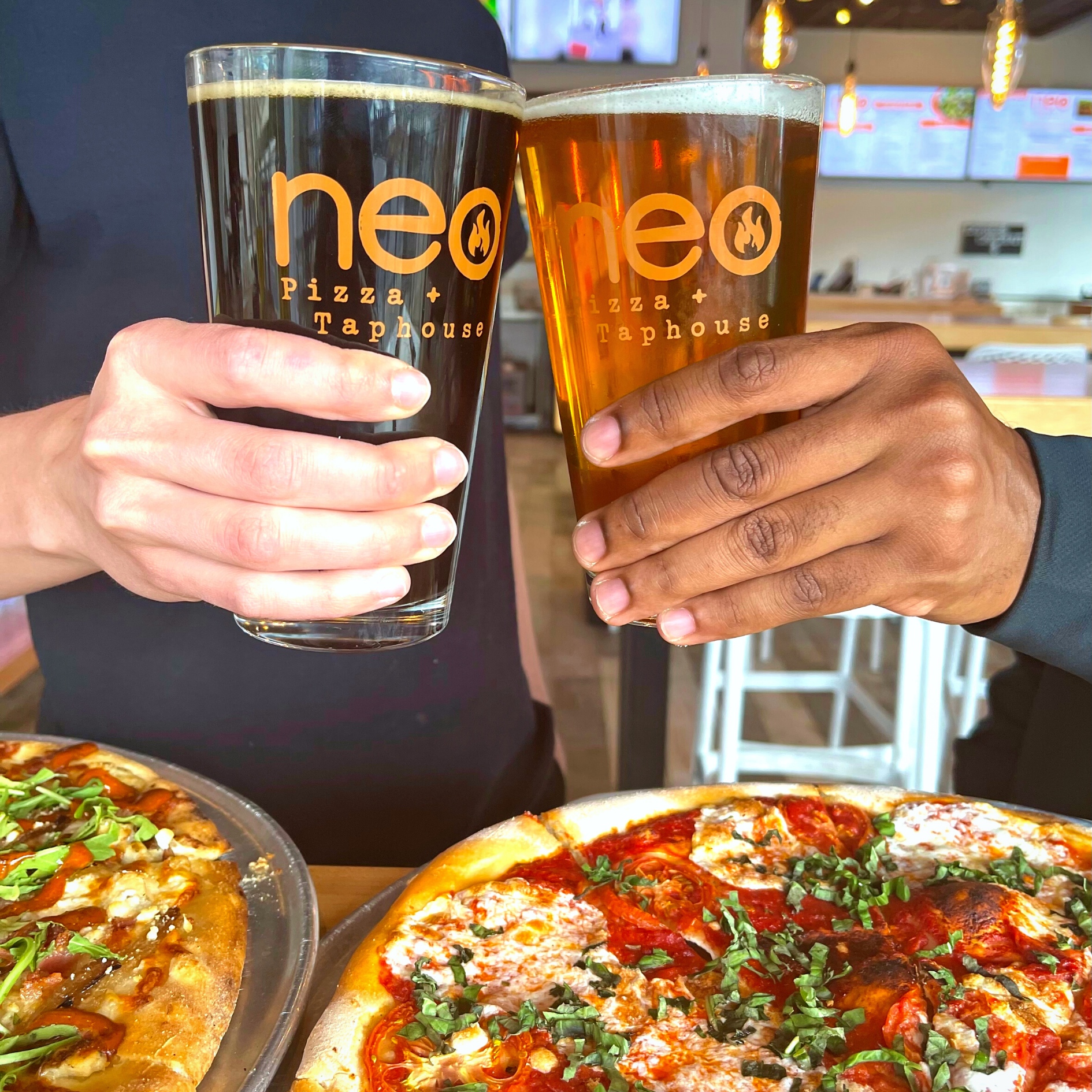 Two people's hands holding glasses of beer with two pizzas on the table below