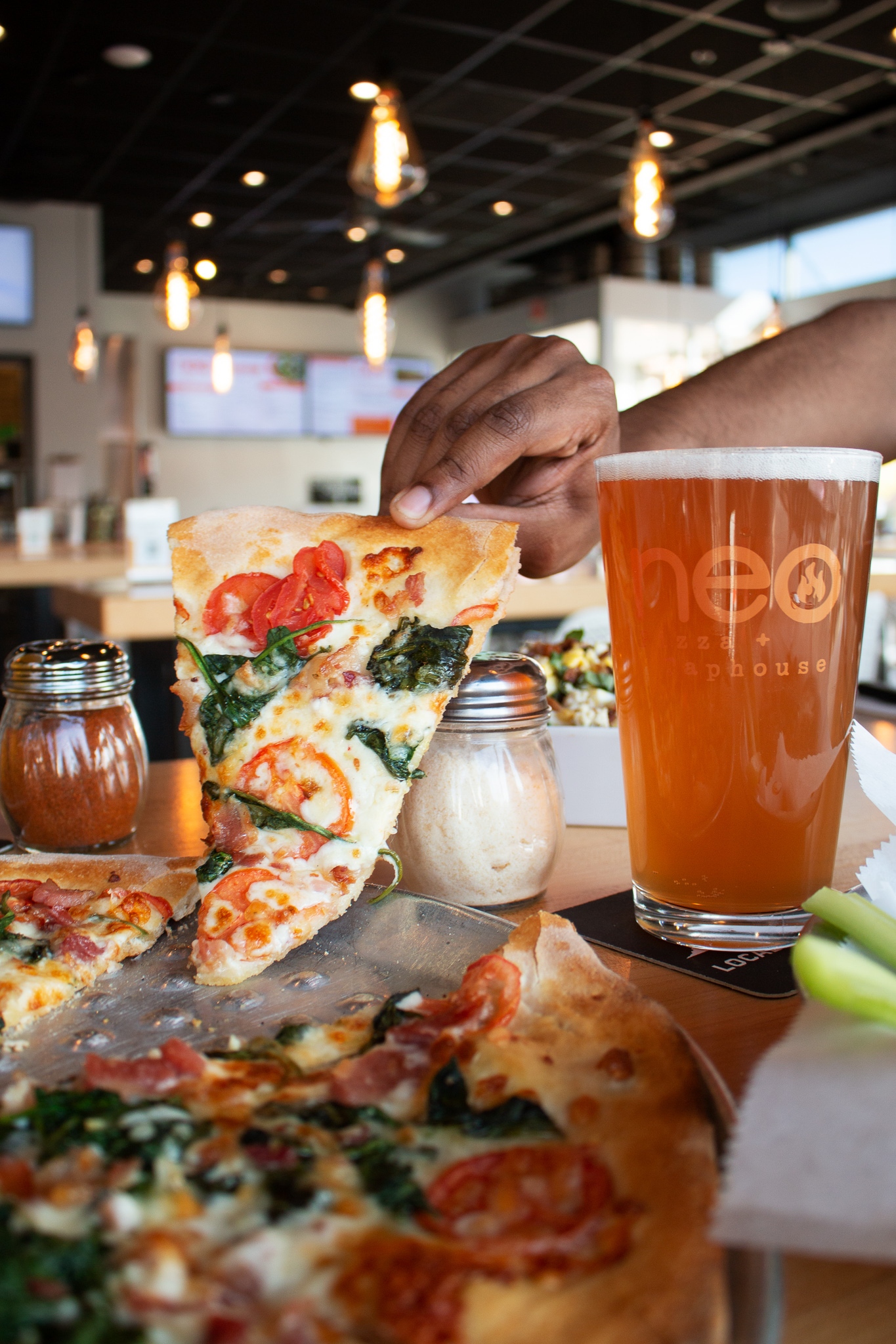 A hand picking up a slice of pizza with a glass of beer on the table