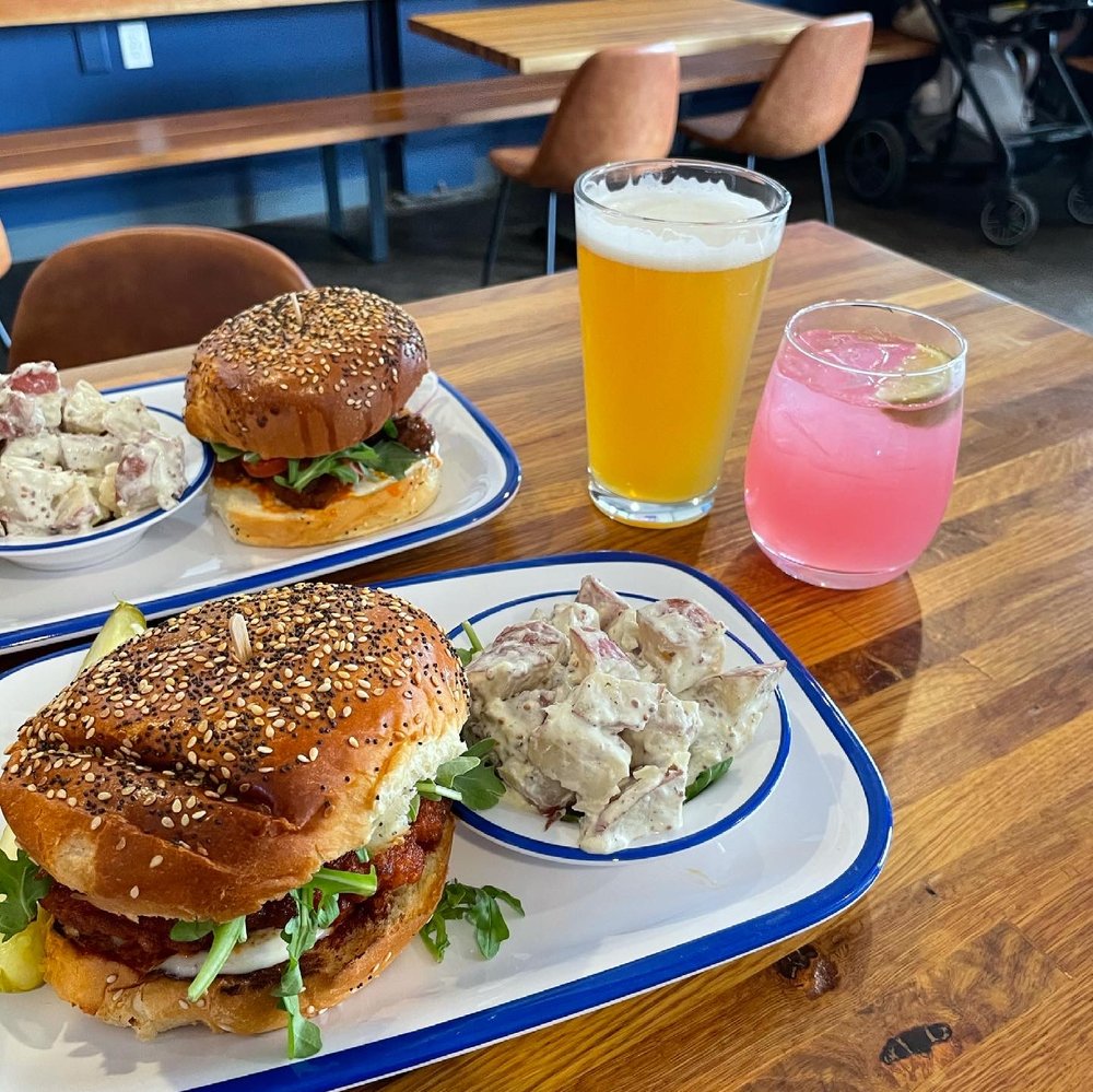 Two plates with a burger and a side, with a glass of beer and a pink cocktail