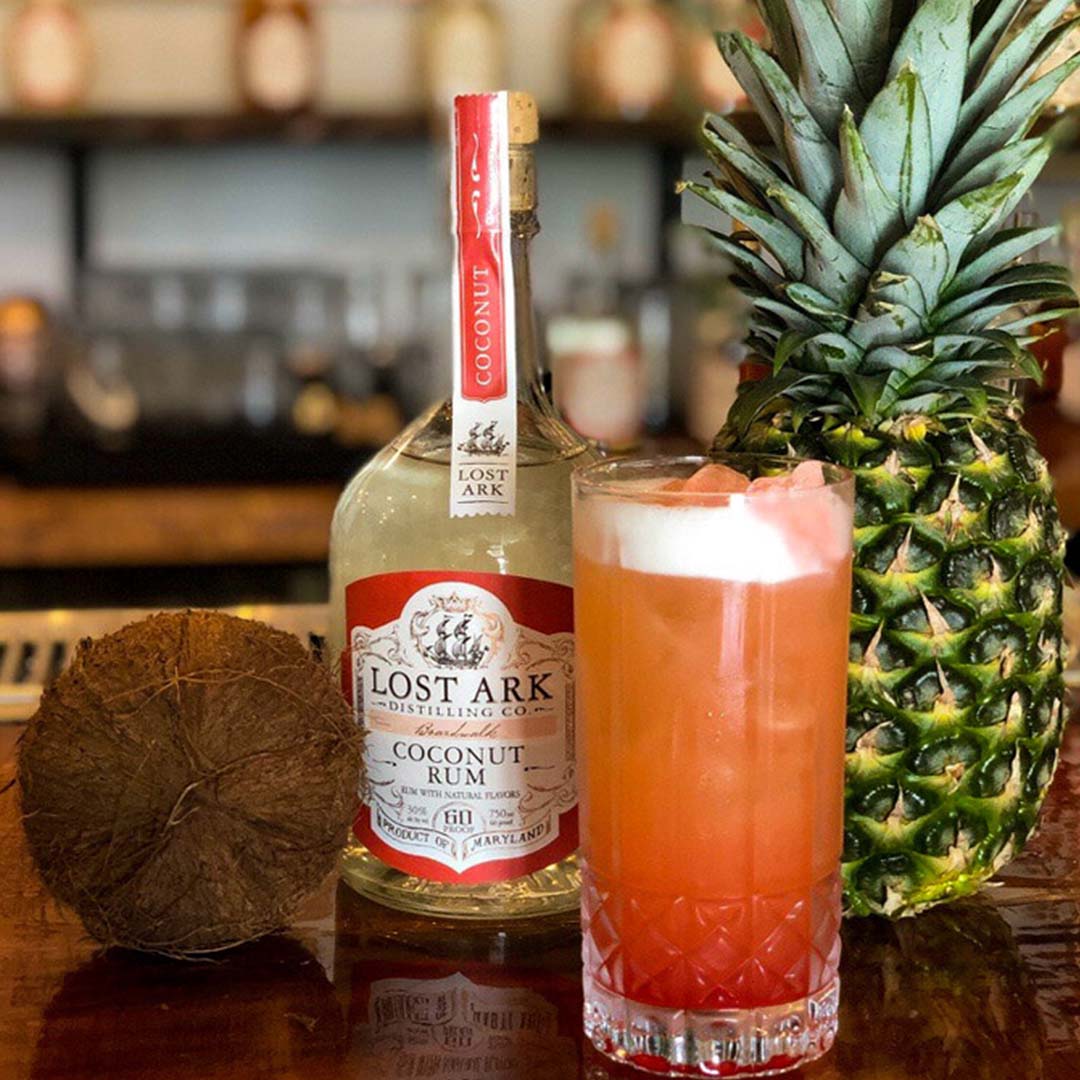 a bottle of coconut rum and a pink orange cocktail, with a pineapple and a coconut on a bar counter