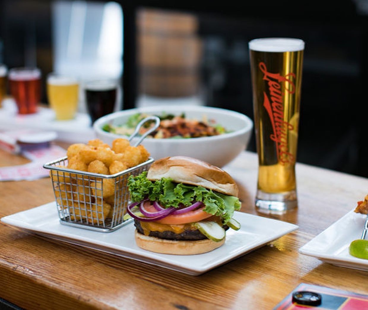 a burger with tater tots, a glass of beer, and other out of focus dishes on a table