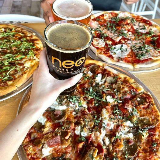 three pizzas on a table with two people cheers-ing glasses of beer