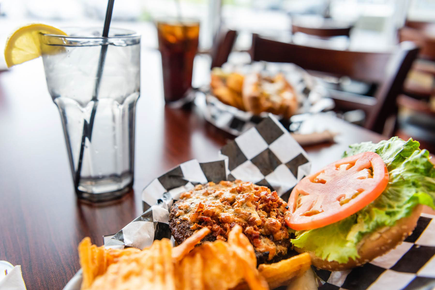 A burger with potato chips and a glass of water with a lemon. Another basket of food and a glass of soda out of focus in the background