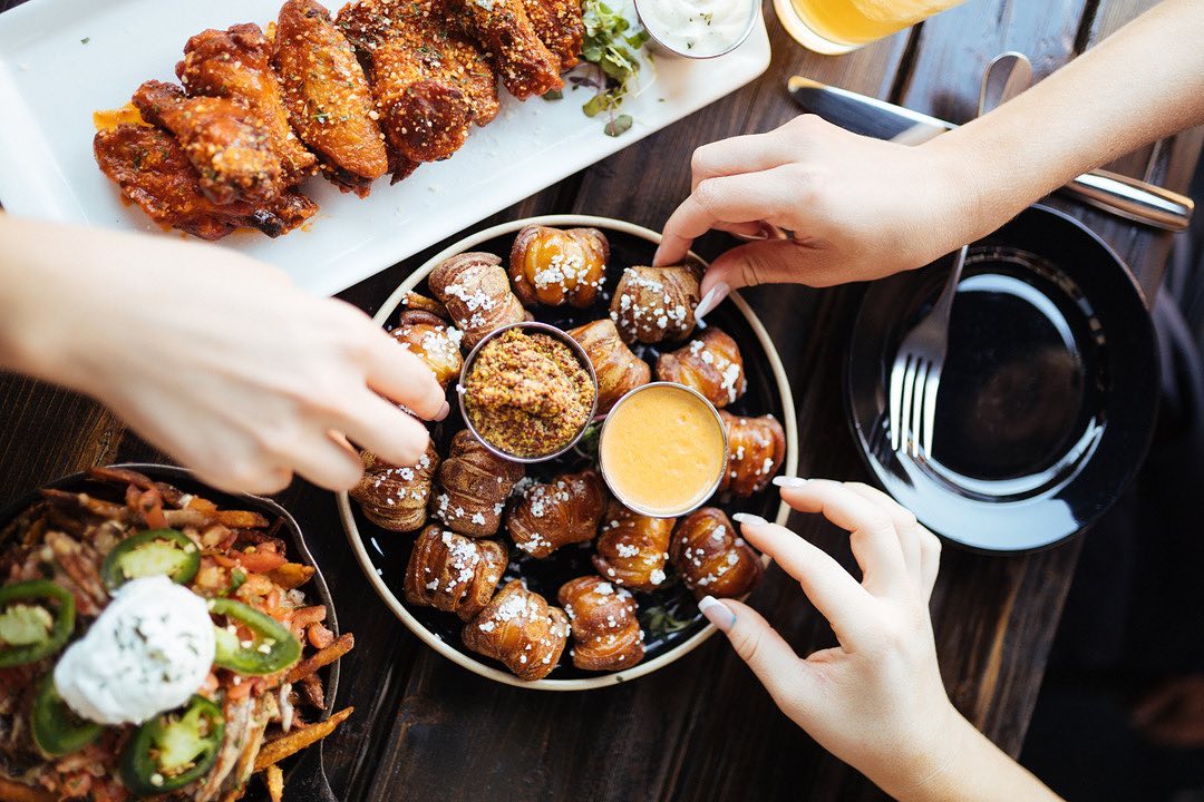 a table with several plates of food, including pretzel bites and wings, with three hands reaching for the food