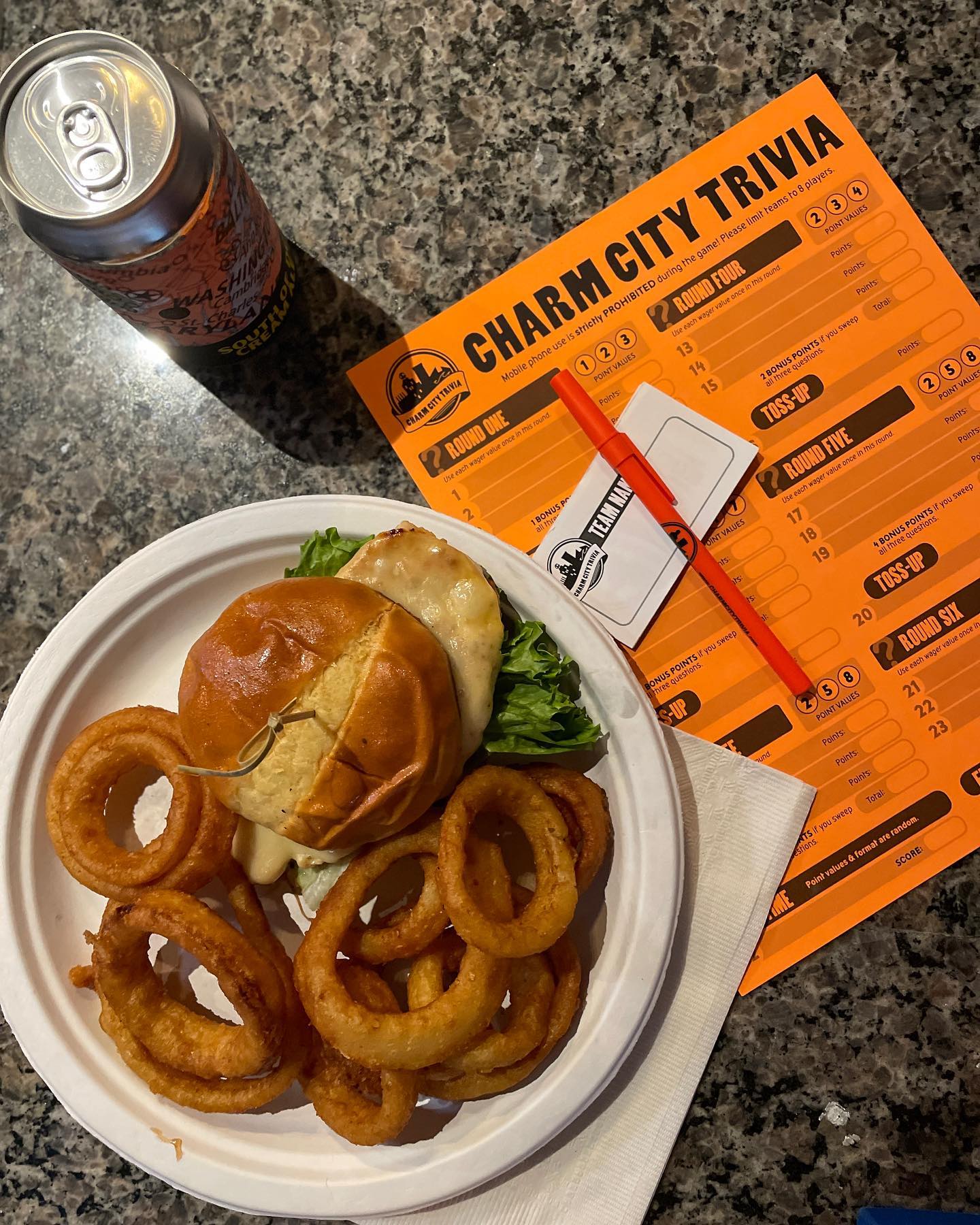 a burger and onion rings on a plate, with a drink and trivia sheet