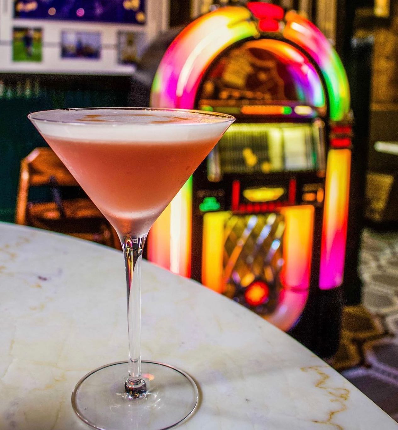 a cocktail on a table with a lit up jukebox in the background