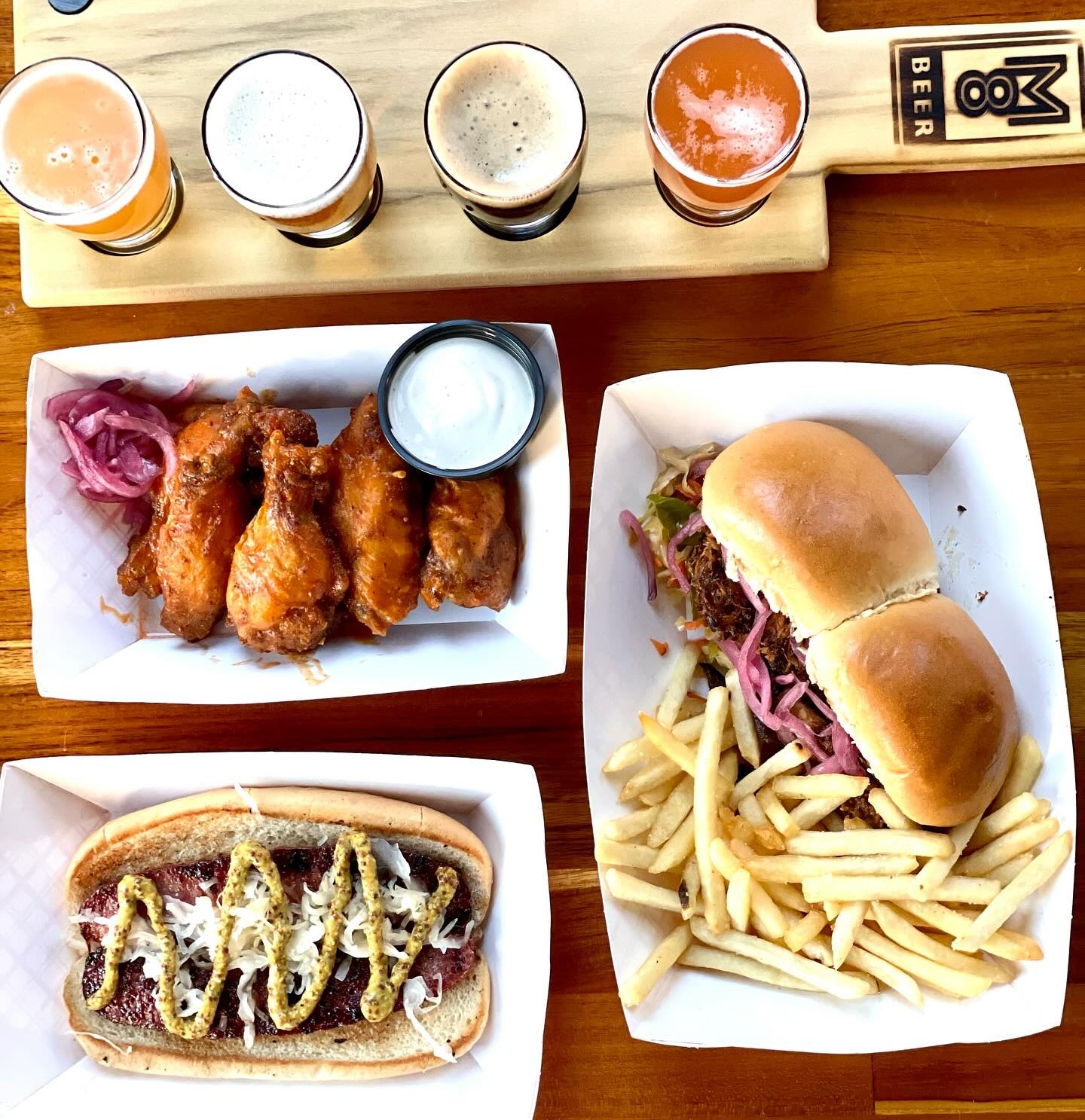 a flight of beer, a plate of wings with red onions and dip, a hot dog, and a sandwich with fries
