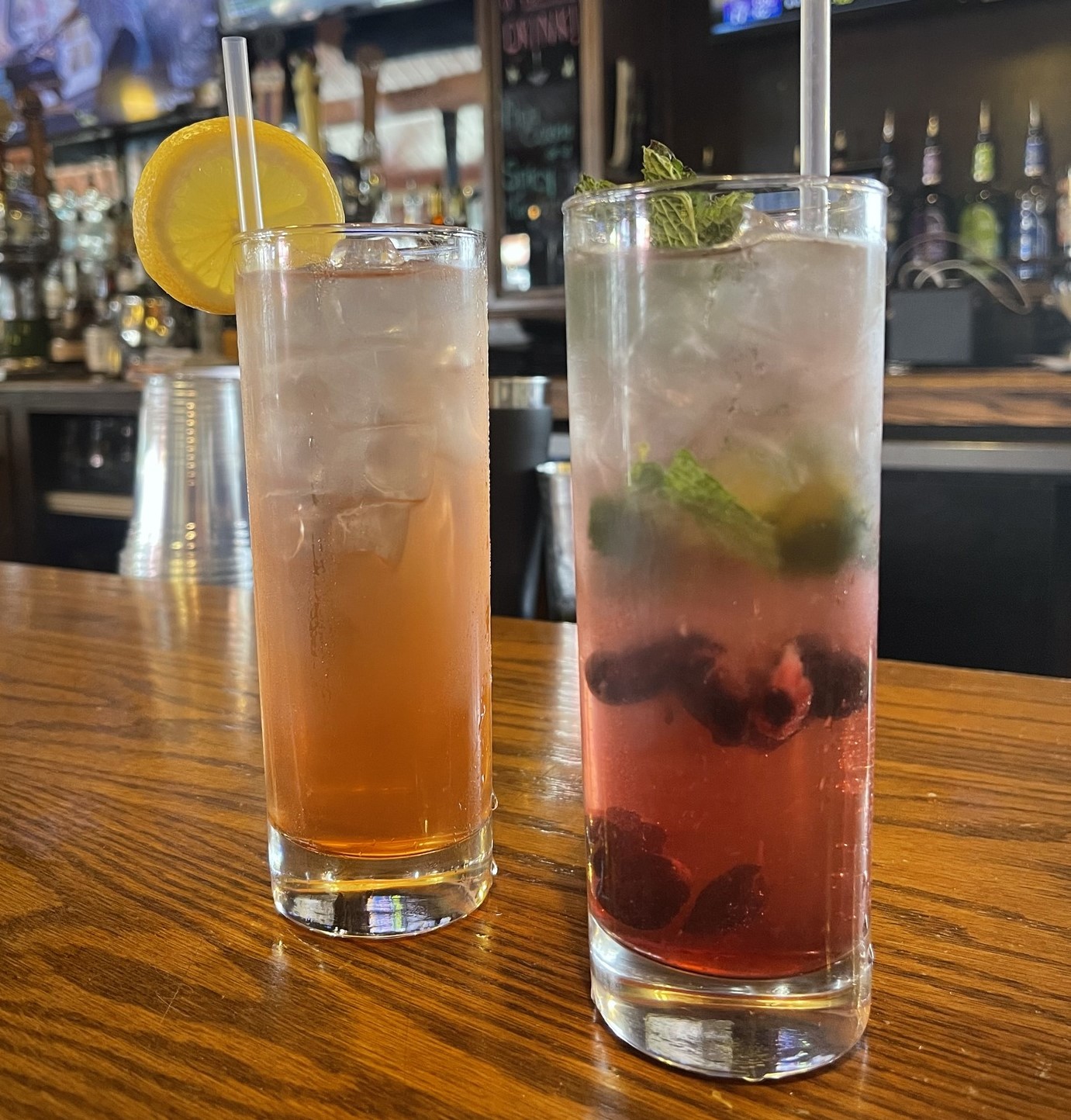 two cocktails, one that's orange colored with a lemon wheel and one that's pink with berries and mint