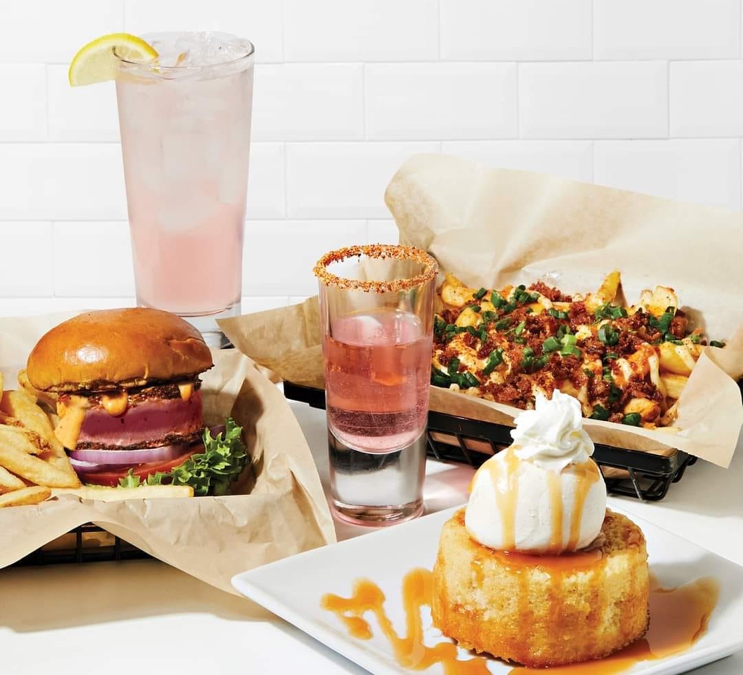a pink cocktail with a lemon wedge, a shot, a caramel lava cake, a tray of cheesy tots, and a burger with fries