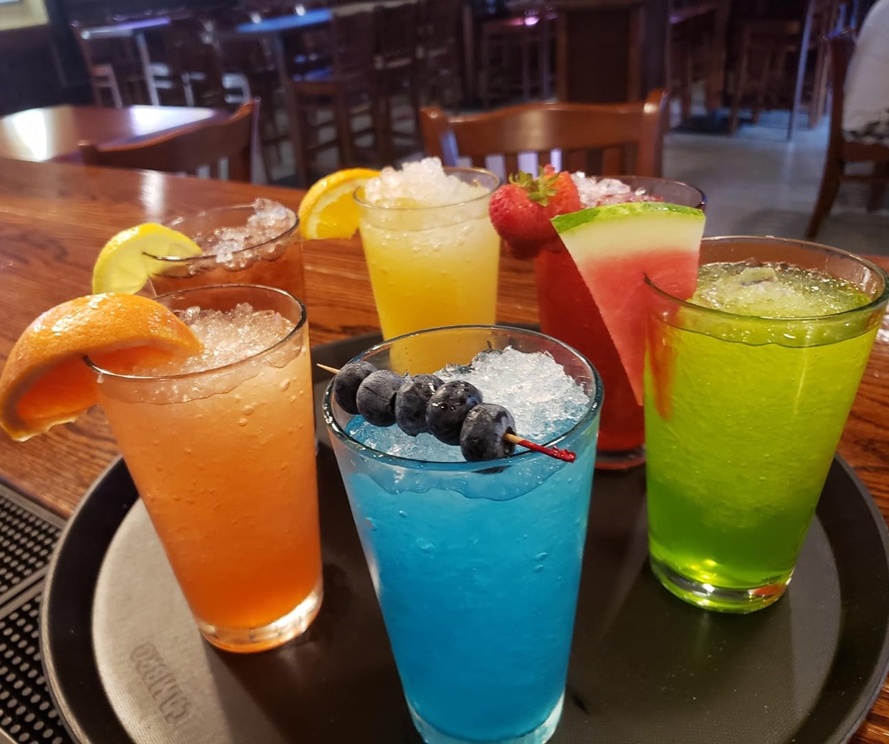 a tray of six colorful drinks, each with a different fruit (including watermelon, blueberries, orange, lemon, grapefruit, and strawberry)on the rim.